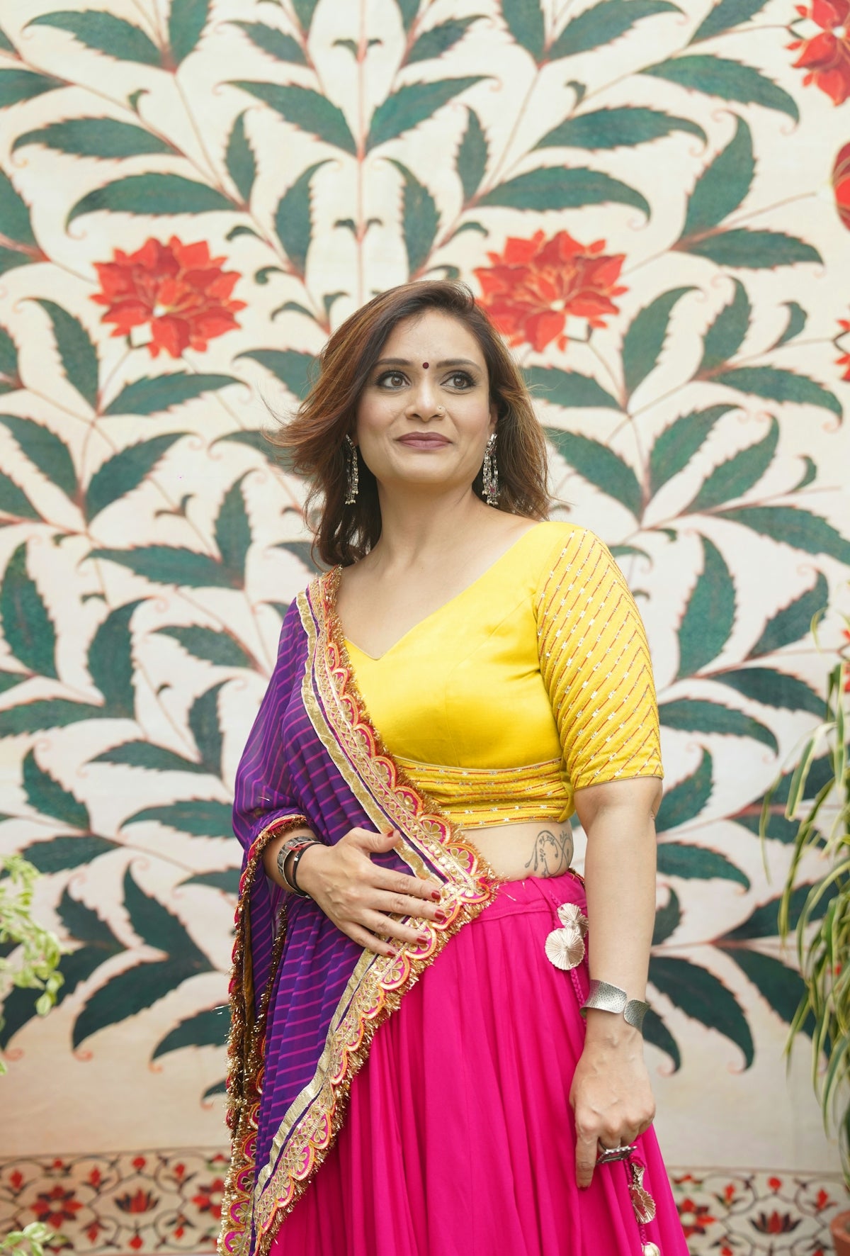 Woman in colorful traditional indian attire poses against floral wall.