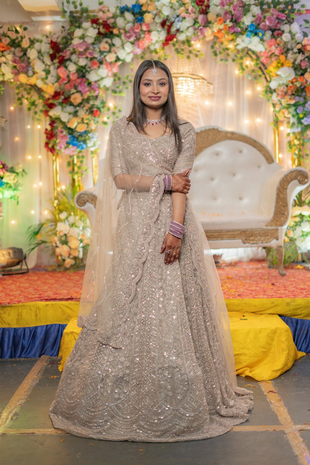 A woman in a sparkling lehenga stands before floral decorations.