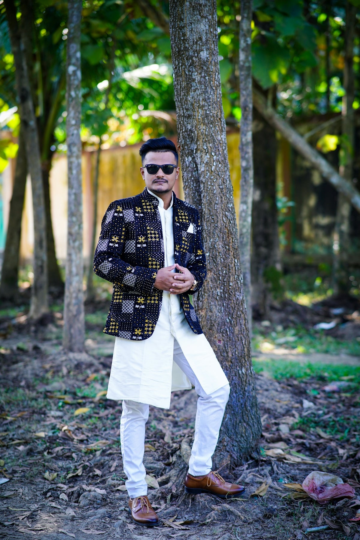Man in patterned jacket and white kurta leaning on tree