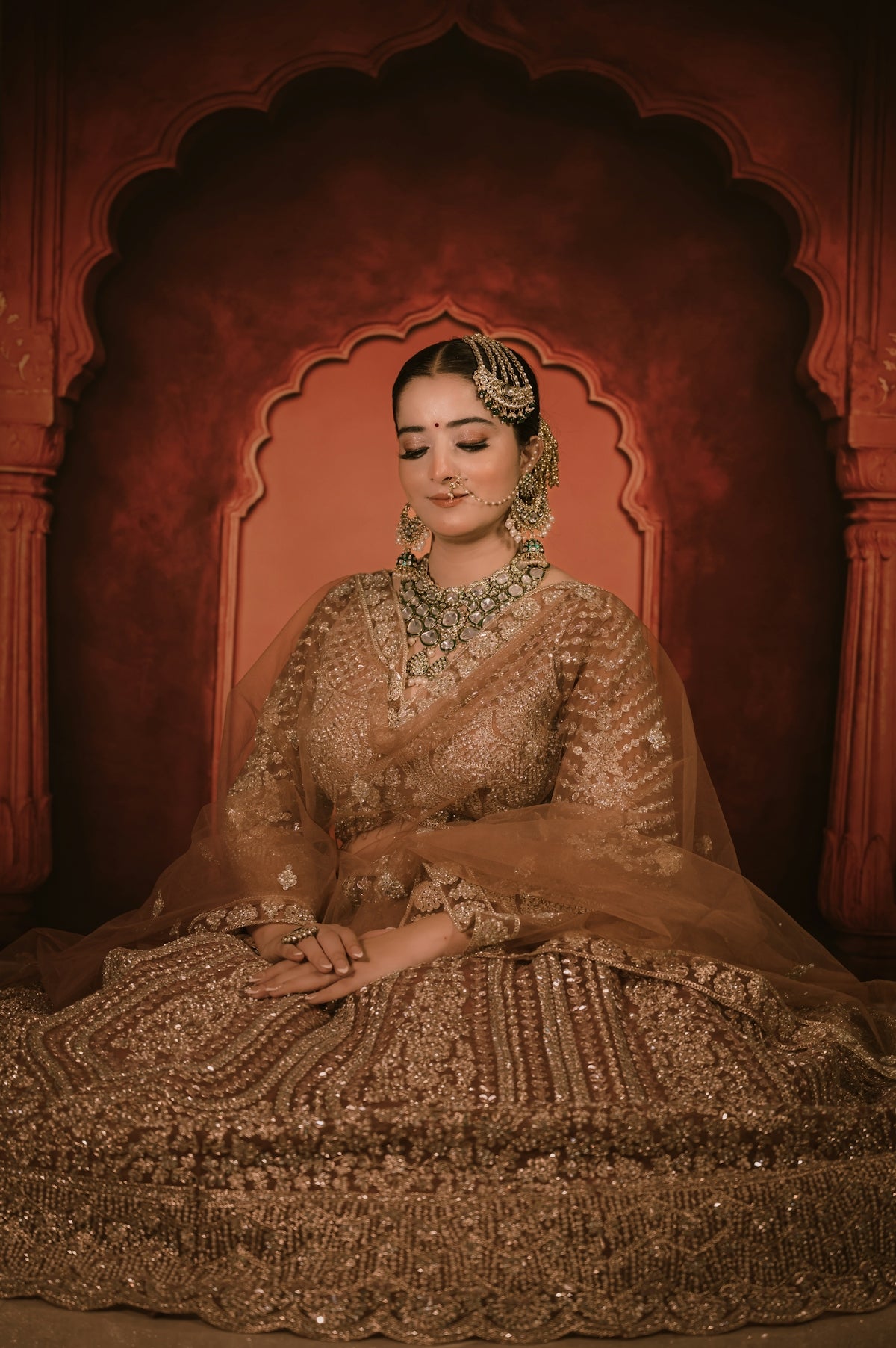 A woman in a gold embroidered lehenga and traditional jewelry against a red archway