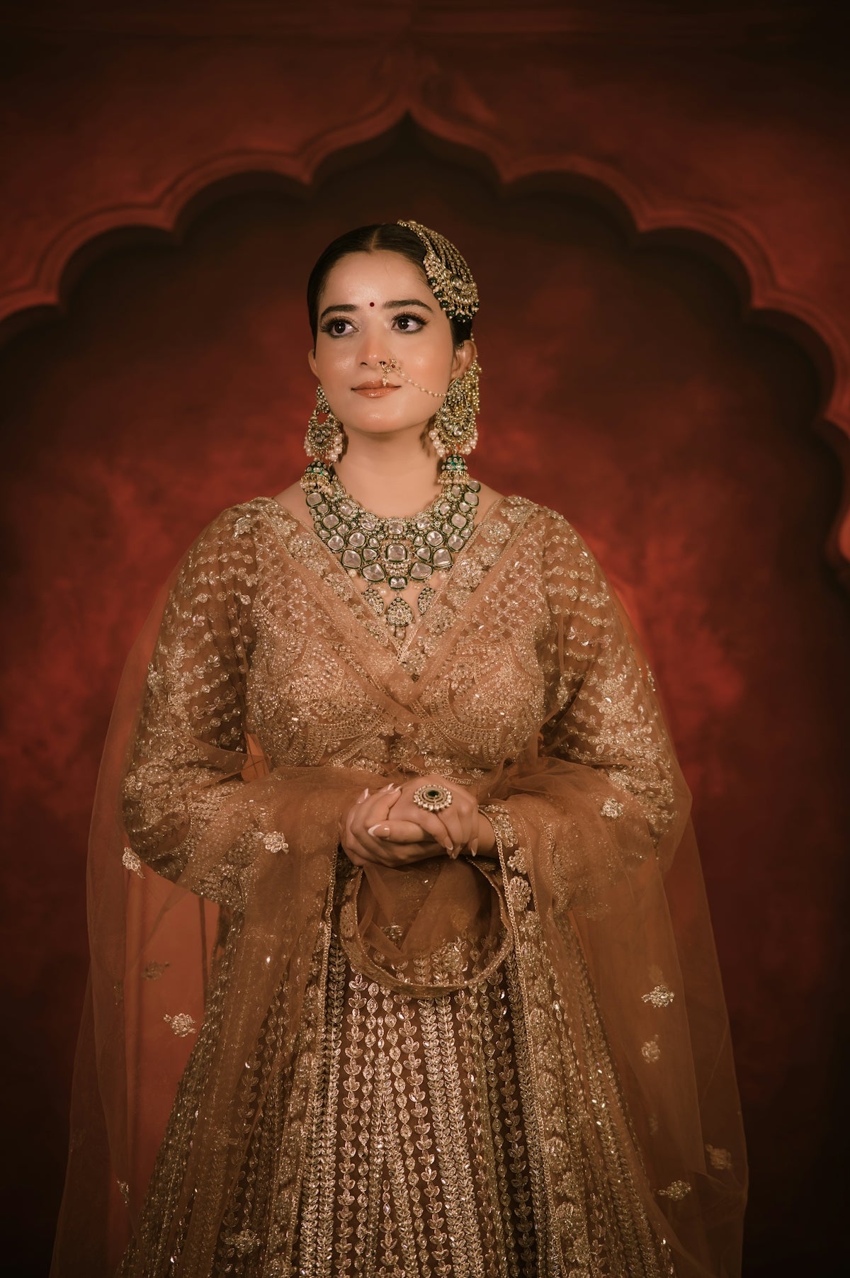 A woman in a gold embroidered lehenga with traditional jewelry against a red background